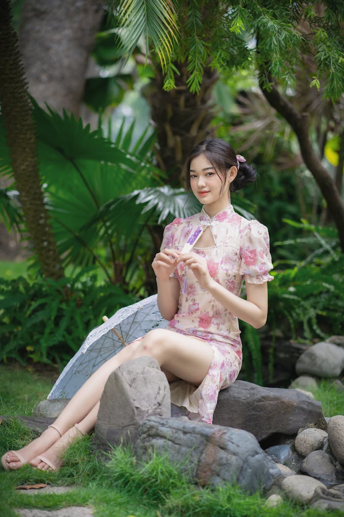 A woman gracefully seated in a lush garden, dressed in a floral cheongsam, holding a fan.