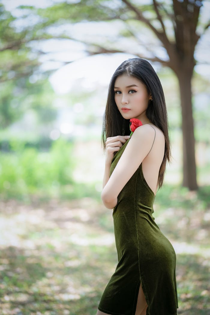 about-img A young woman in a green dress holding a red rose in a serene outdoor setting.